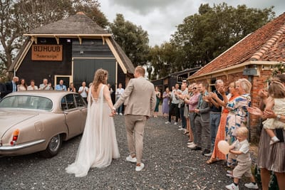 Thijs en Lois lopen hand in hand tussen twee rijen applaudisserende gasten, een klassieke auto naast hen