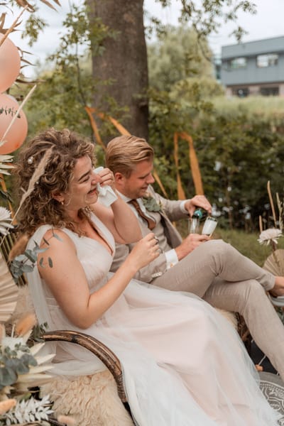 Lois droogt lachend haar tranen terwijl Thijs champagne inschenkt op hun bruiloft in het groen