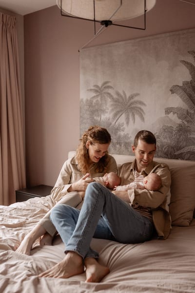 Vader en moeder zitten op bed en houden elk één van de tweelingzusjes Veerle en Lola vast, voor behang met palmen