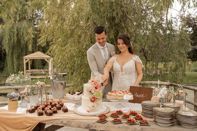 Dominique en Daimy snijden samen de bruidstaart aan bij een tafel vol gebakjes in de tuin