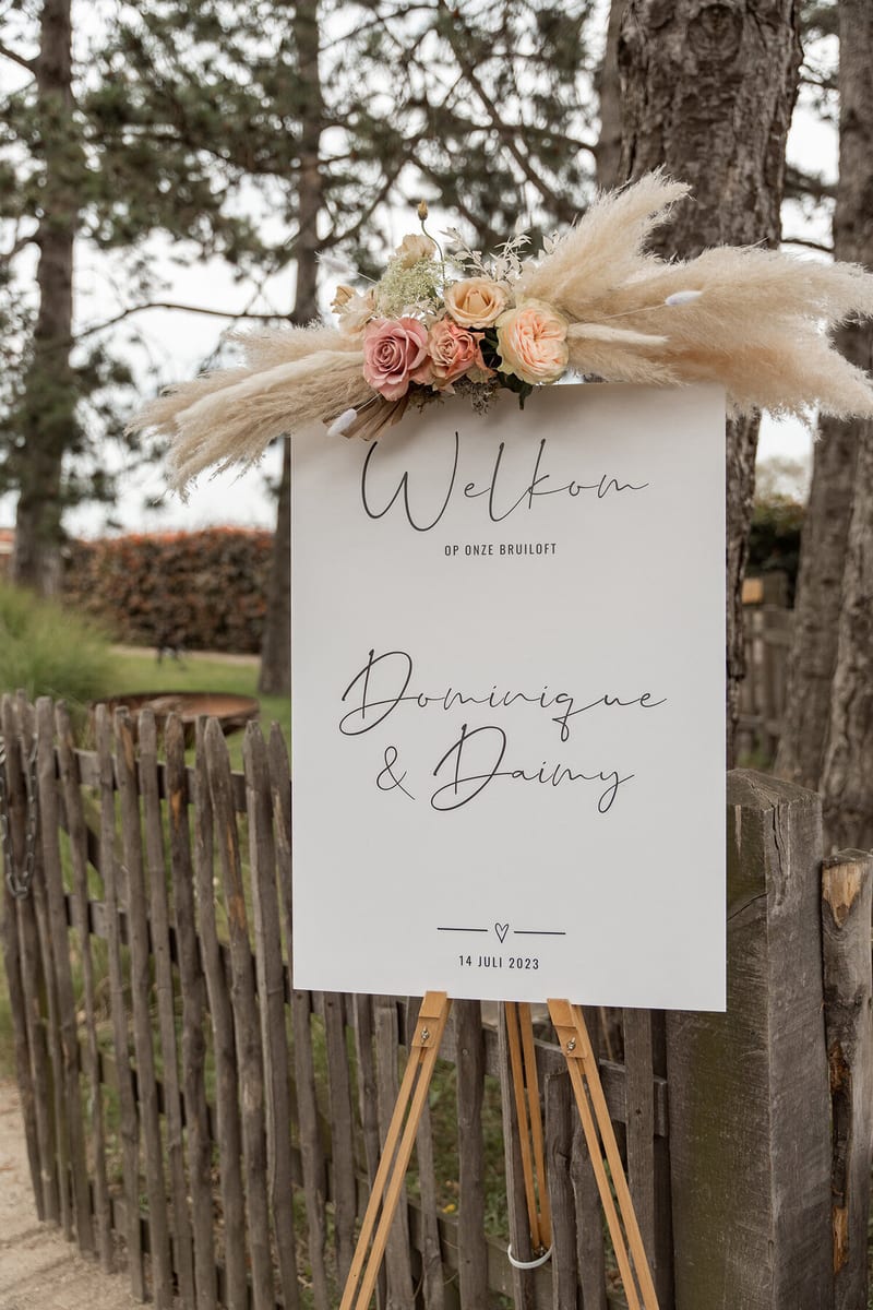 Welkomstbord met pampaspluimen en rozen kondigt de bruiloft van Dominique en Daimy aan bij een houten hekje