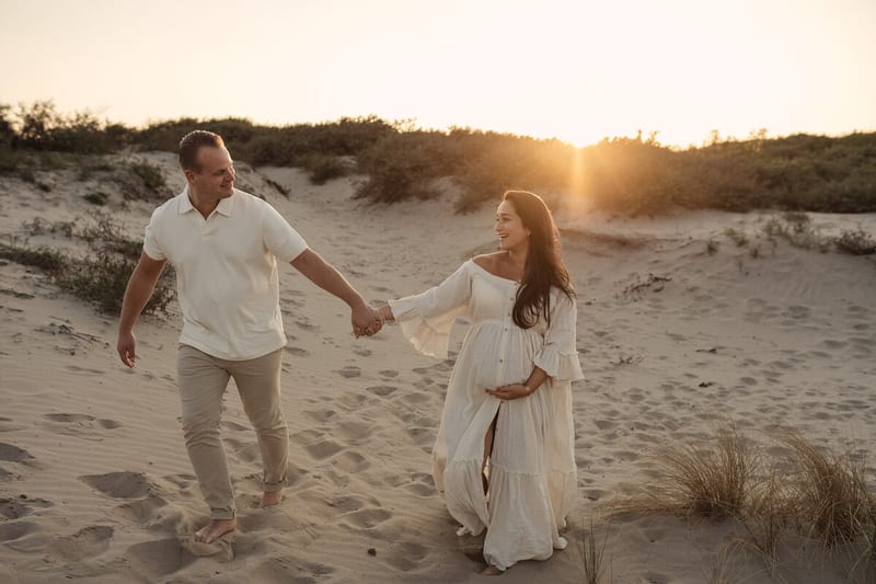 Zwanger stel loopt hand in hand door het mulle duinzand terwijl de zon laag achter de duinen zakt