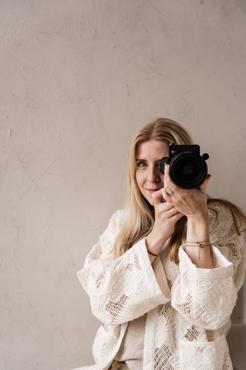 Romy van Leeuwen richt haar camera op de kijker, half verscholen achter de zoeker tegen een lichte pleisterwand