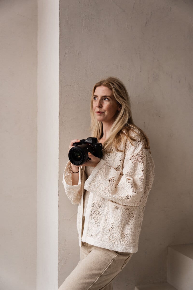 Romy van Leeuwen staat met haar camera tegen een lichte wand en kijkt opzij in zacht daglicht