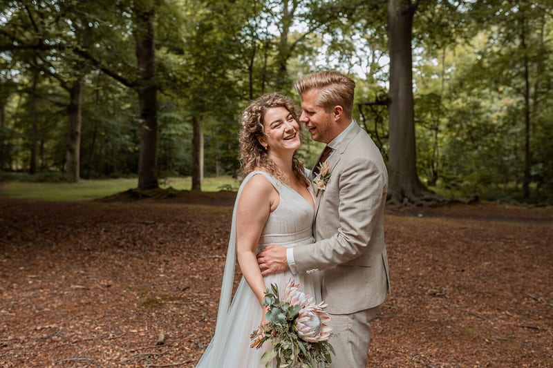Thijs en Lois staan lachend wang aan wang in het bos, een boeket met grote protea in haar hand